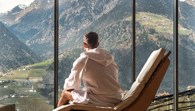 15 Erholungsorte, die Stil und Zeitgeist atmen 3 Mann im Bademantel schaut aus dem Fenster im Wellnessbereich auf ein Alpenpanorama
