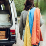 3 Outdoor-Textilien für den Alpenausflug 7 Person hat drei Handtücher auf dem Rücken und steht vor der geöffneten Heckklappe seines Campervans