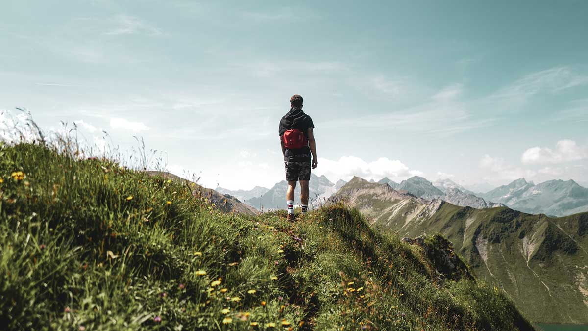 Für den frühen Wurm: Frühlingsurlaub in den Alpen 2026 2 Ein junger Mann mit Rucksack und Wanderschuhen blickt auf eine Frühlingswiese in den Alpen