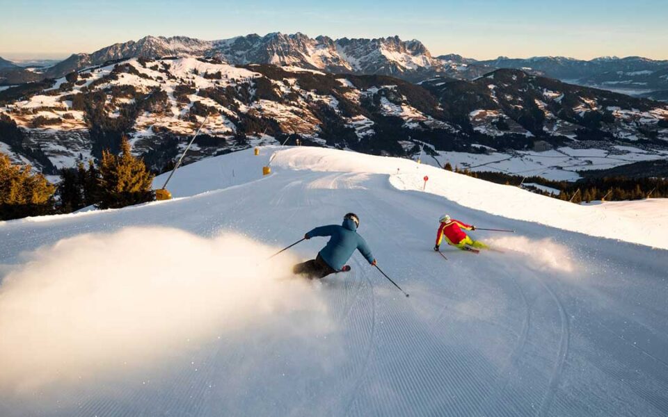 Zwei Menschen sausen mit Skier die Piste in den Alpen im Wilden Kaiser herunter, und sind voller Vorfreude auf den nächsten Frühlingsurlaub